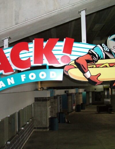Bold sign reading 'SACK! FAN FOOD' in a stadium, featuring a football player holding a giant hot dog.