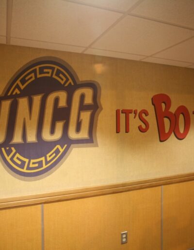 UNCG logo and text reading "IT'S BO TIME!" on a textured wall in a well-lit corridor.