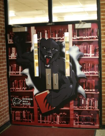 A large black bear bursts through a library door, surrounded by colorful bookshelves.