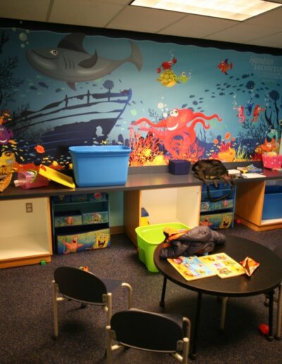 Playroom with colorful underwater mural, toys, and a child watching TV on a small cart.