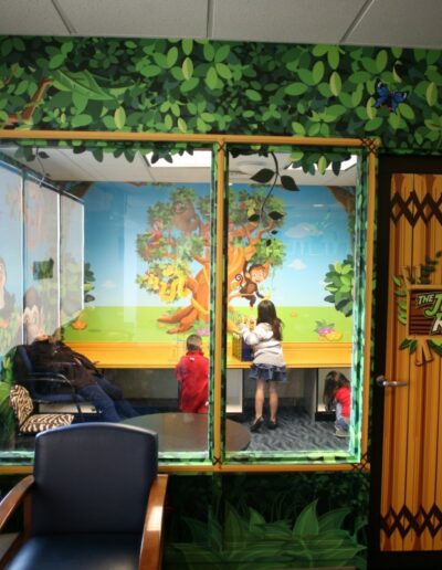 A colorful room with jungle-themed murals and a "Jungle Room" sign. Inside, children play under a tree painting.